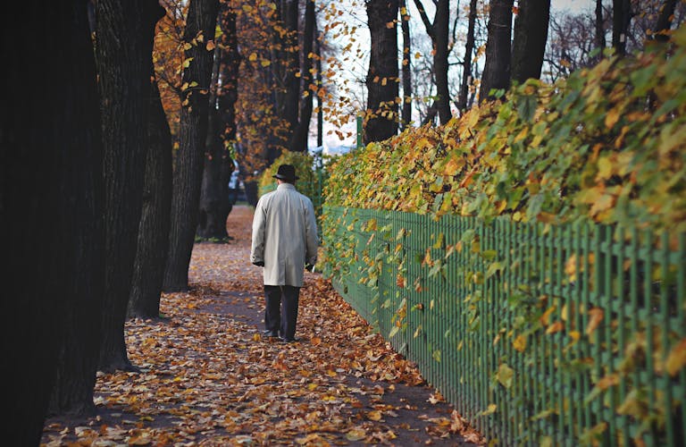 A man in a coat and hat walks along a leaf-covered path beside a fence in an autumn park.